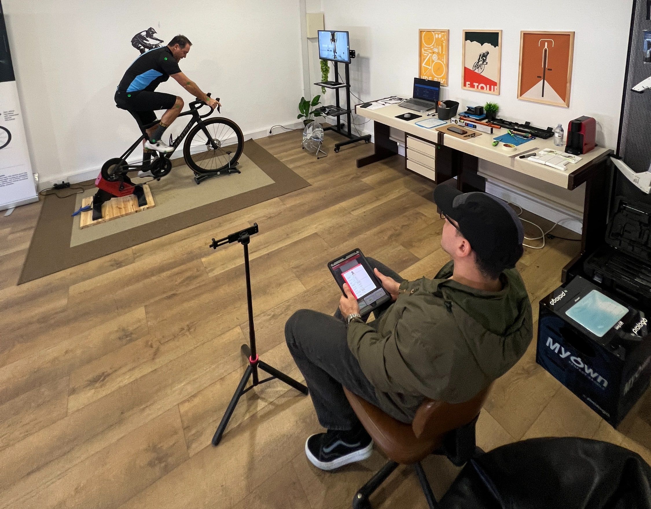 Sessão de bike fit no estúdio VTR Bike Lab, em Oeiras: ciclista a pedalar no rolo, Victor sentado com o iPad e a análise da pedalada no ecrã da televisão